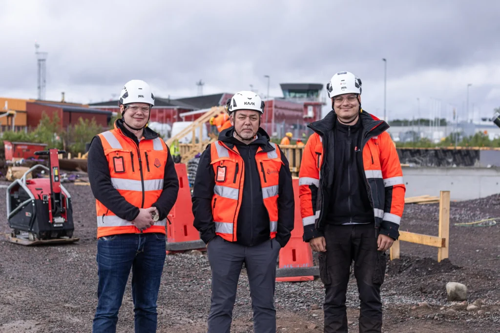 Tre av Anfras medarbetare står bredvid varandra och ler mot kameran på byggarbetsplatsen. De bär lämplig skyddsutrustning, såsom orangea varselkläder och hjälmar. I bakgrunden skymtar flygplatsens byggnader.