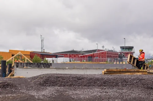 En byggnadsarbetare klädd i varselkläder flyttar en gjutform i trä bredvid en lång betonggrund. I bakgrunden syns Kiruna flygplats flygledartorn och röda byggnader.