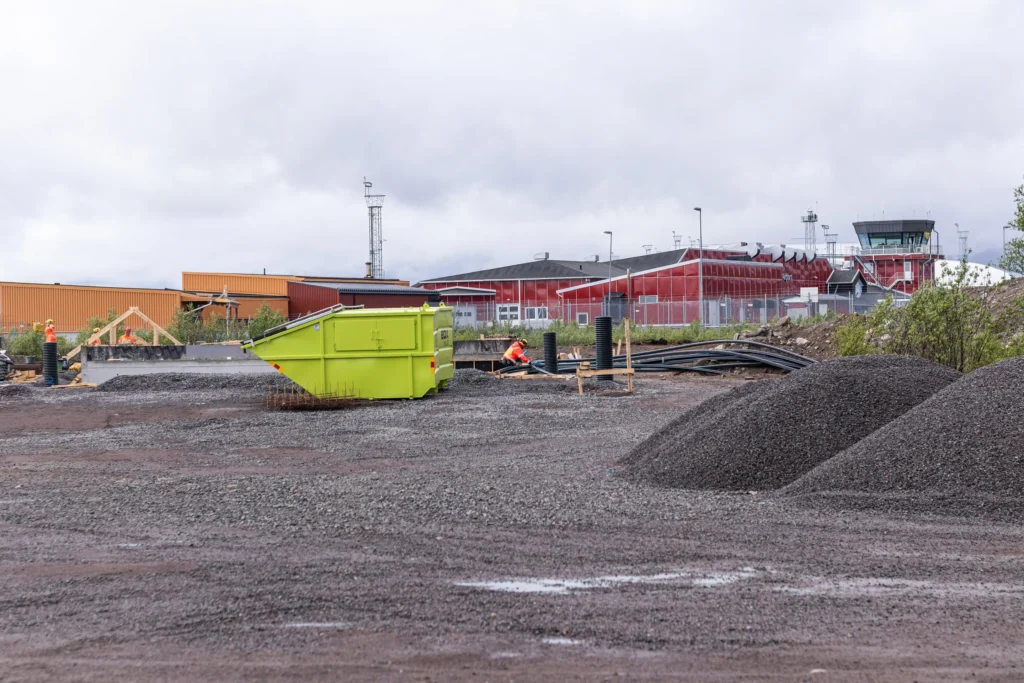 En pågående byggarbetsplats med grus, en grön container och byggmaterial. I bakgrunden syns Kiruna flygplats röda byggnad och flygledartornet.